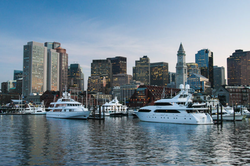 A view of the Boston skyline and Harbour taken from a boat at sunset. Yachts are visible in the foreground.