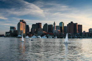 A view of the Boston skyline and Harbour taken from a boat at sunset. Boats are sailing on the water in the foreground.